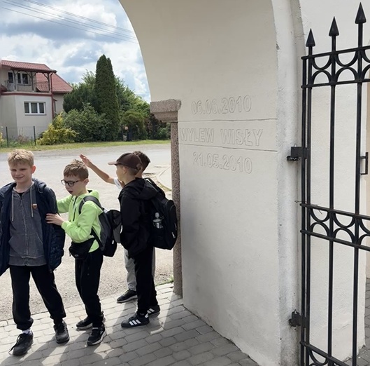 Czwórka dzieci stoi przy białym murze z datą i czarną metalową bramą, na podwórku.