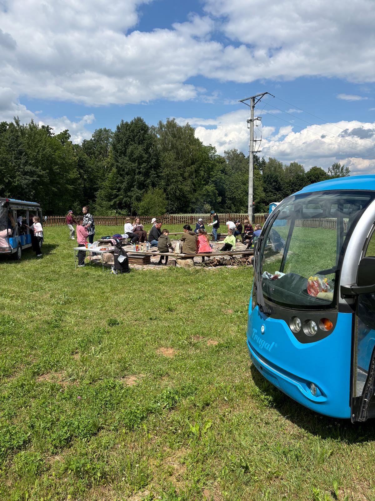 Niebieski Ekobusik pojazd elektryczny stoi na trawie, a dzieci siedzą przy stole piknikowym przy ognisku.