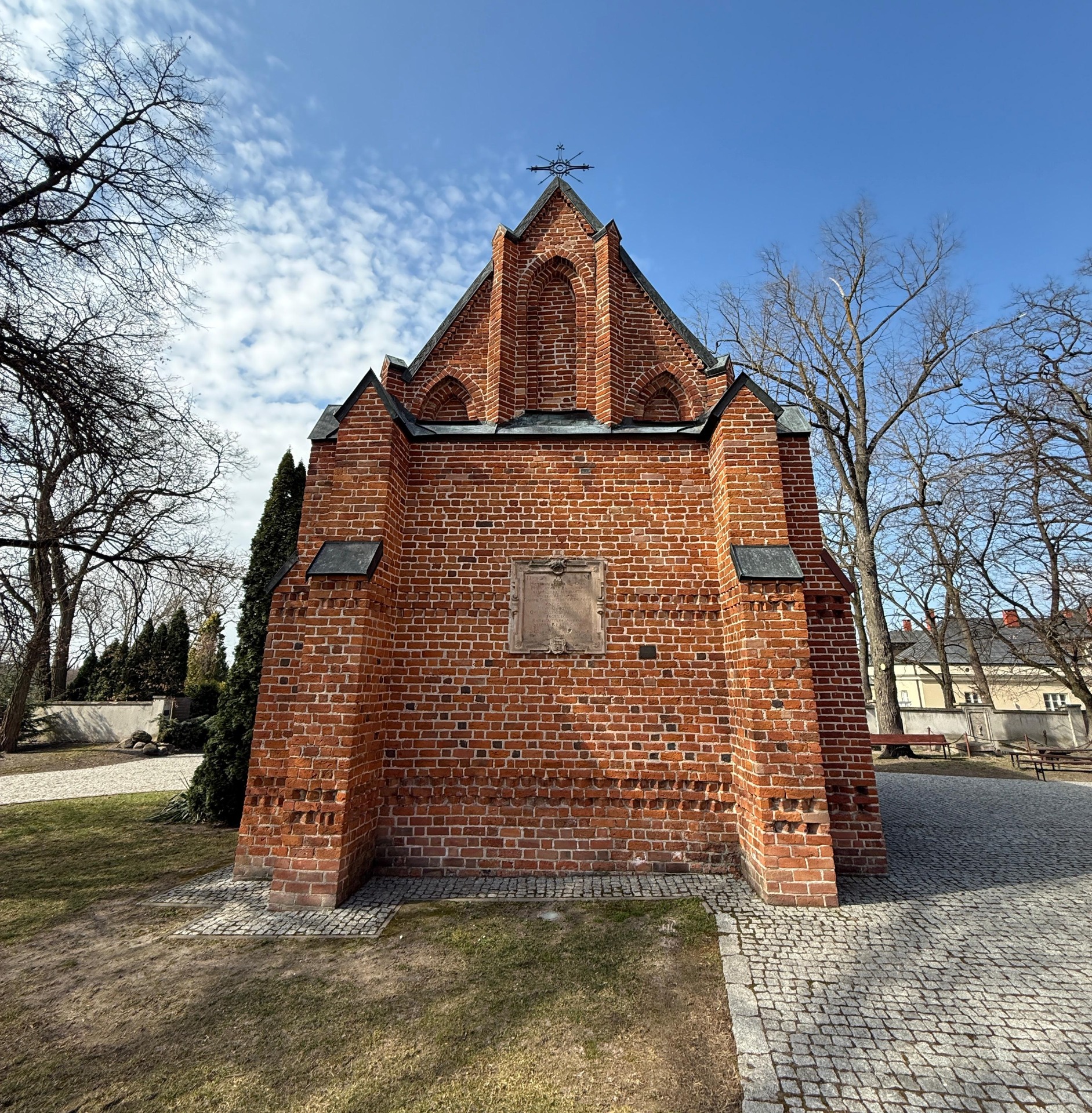 Kościół św. Stanisława Biskupa i św. Tomasza Apostoła w Piotrawinie, Ceglany budynek z trójkątnym szczytem, łukowymi wnękami i metalowym krzyżem na tle nieba i drzew.